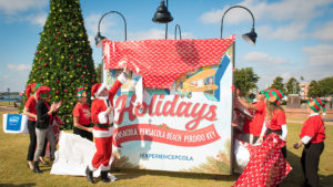 People dressed as santa and christmas elves near a large christmas tree unveiling a sign that says, "Holidays - Pensacola, Pensacola Beach, Perdido Key. #EXPERIENCEPCOLA"